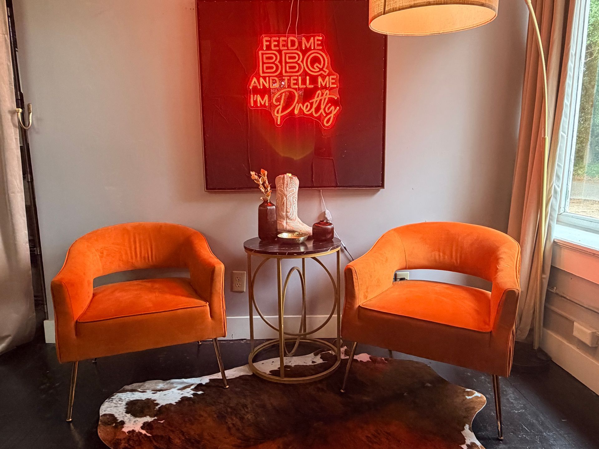 Cozy seating nook with two orange chairs, round table, floor lamp, and cowhide rug beneath wall art.