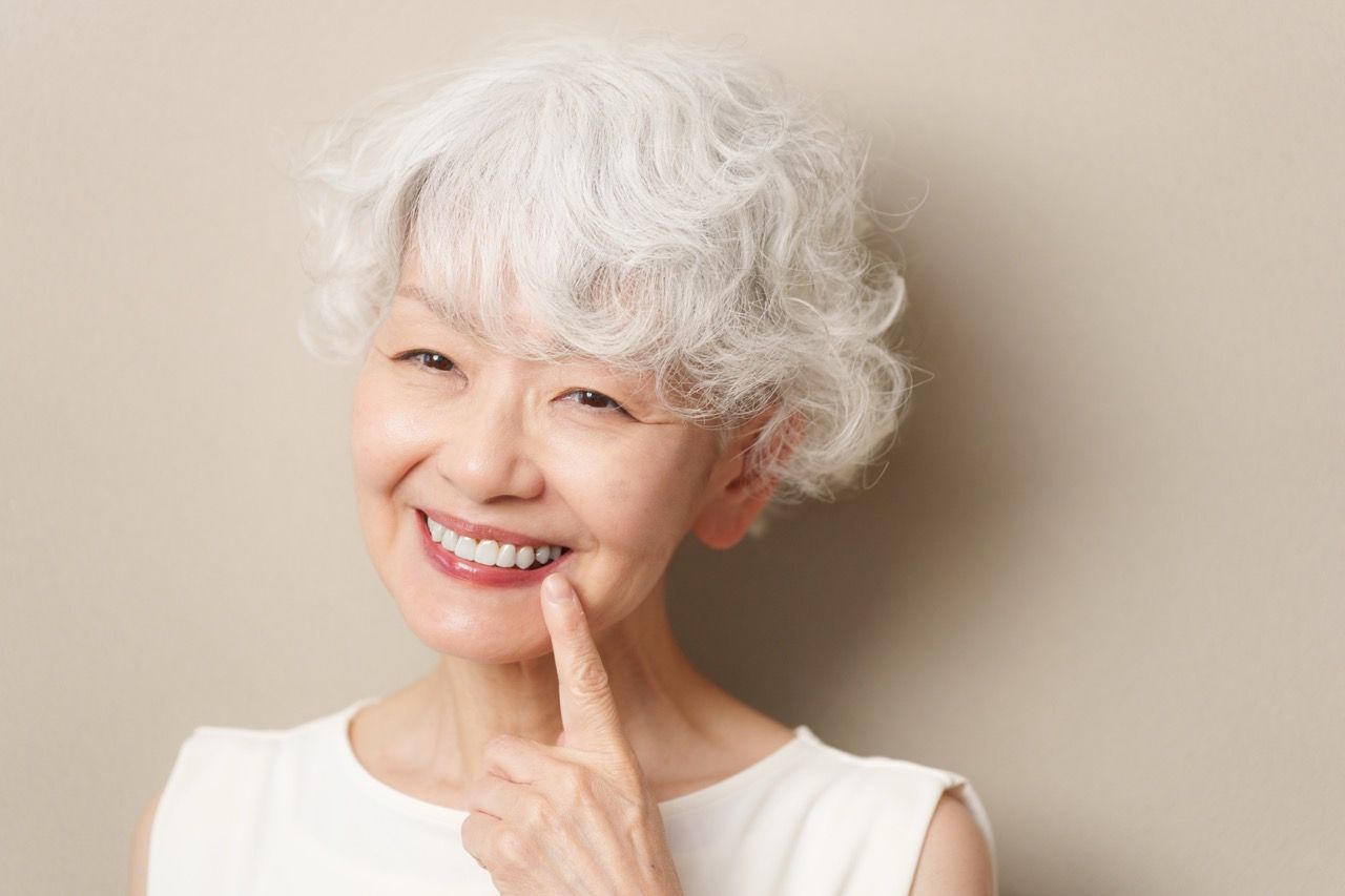 Smiling woman with white curly hair touching her chin against a beige background