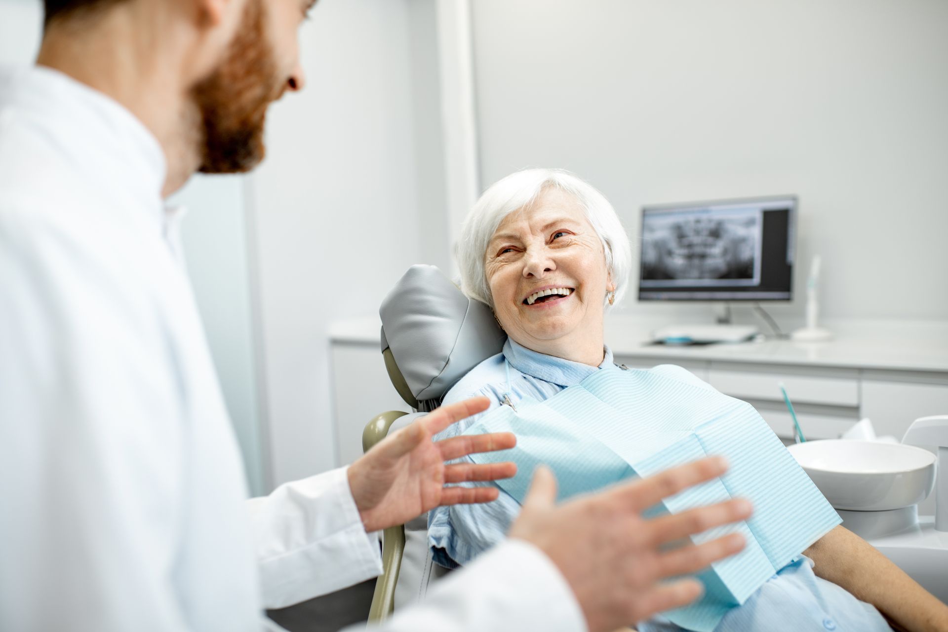 Smiling senior woman with white hair sitting in a dental chair, talking with a dentist in a white coat, with a dental X-ray displayed on a screen in the background.