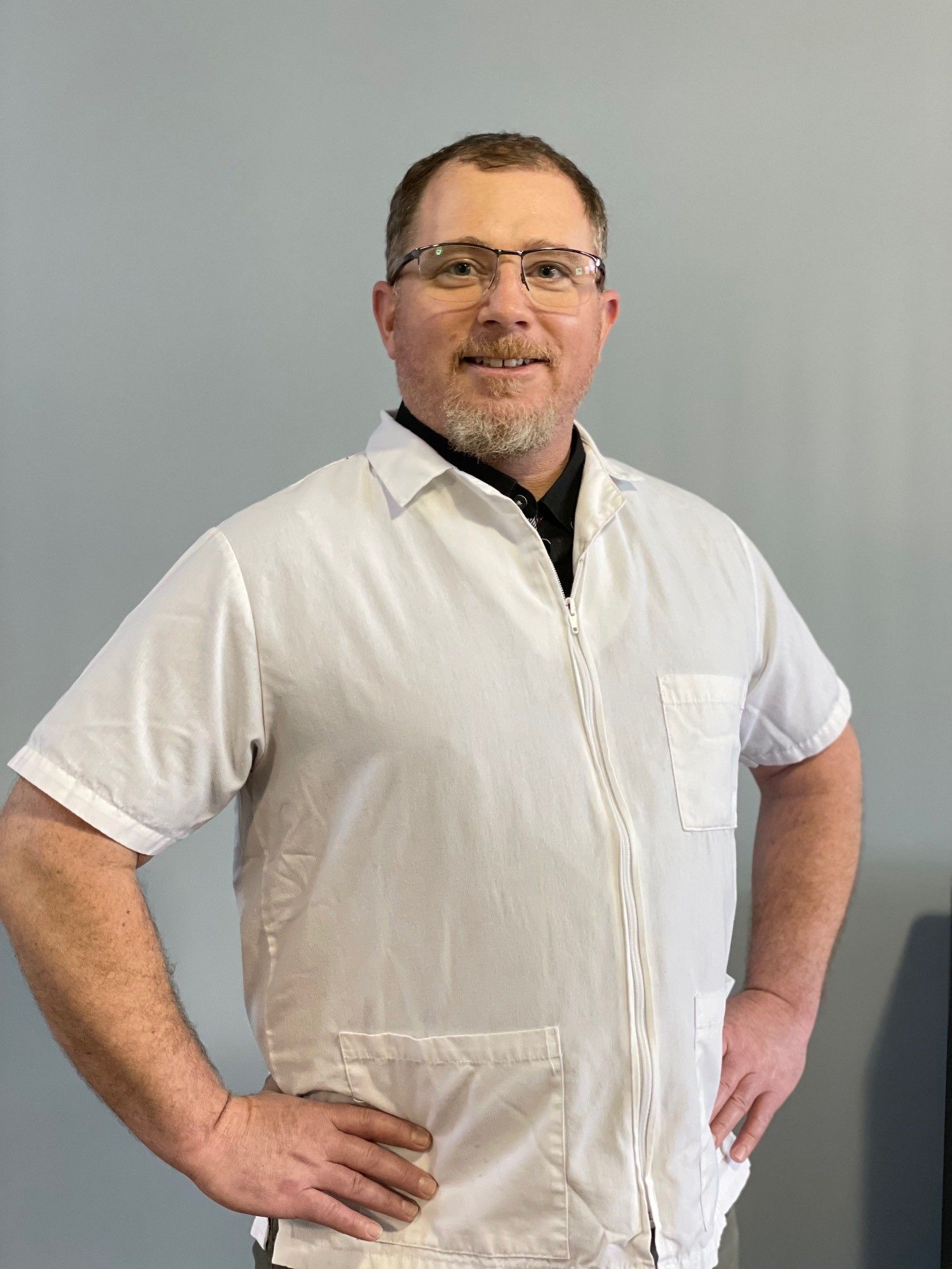 Smiling man with glasses and a short beard wearing a white clinical jacket, standing with his hands on his hips against a gray background.