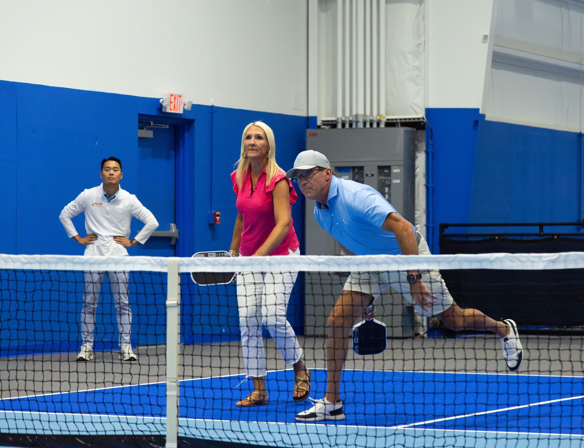 Three pickleball players on a court; a man swings, woman prepares, and other watches.