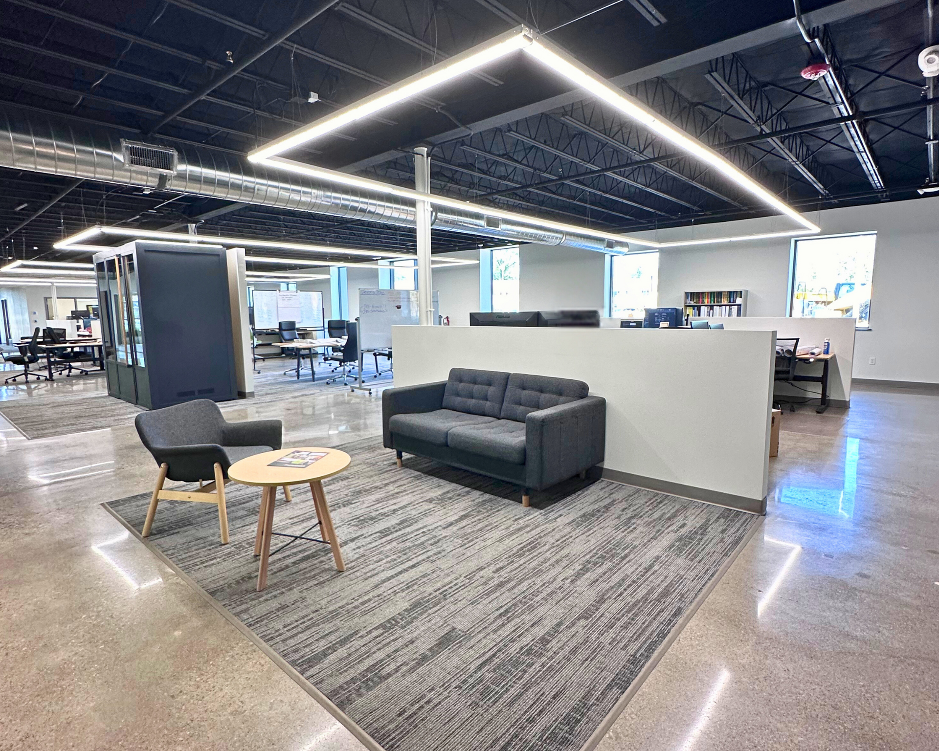 Modern office space with seating area: dark sofa, chair, round table on a rug. Cubicle area in the background.