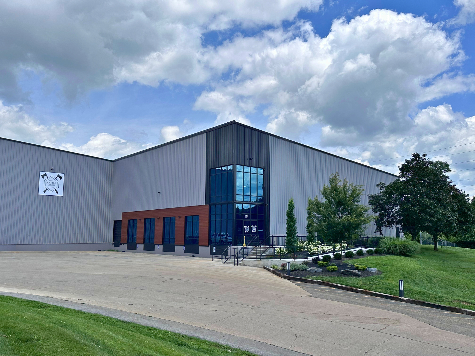 A large industrial building with a glass entrance. Gray and brown exterior, green lawn, cloudy sky.