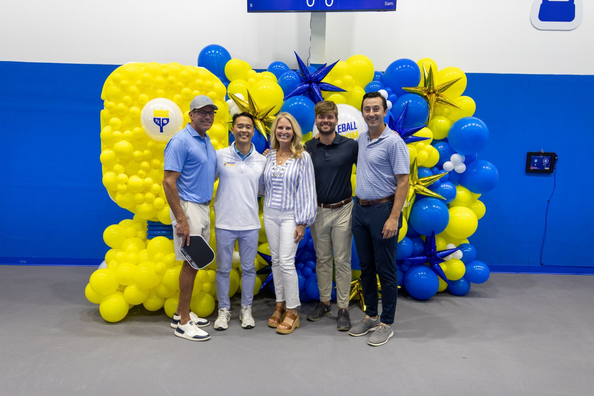 Five people pose in front of a blue and yellow balloon backdrop inside a building.