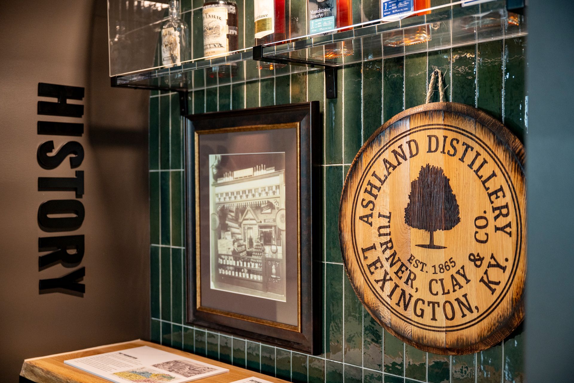 Ashland Distillery historical display: framed image, wooden sign, green tile backdrop, 