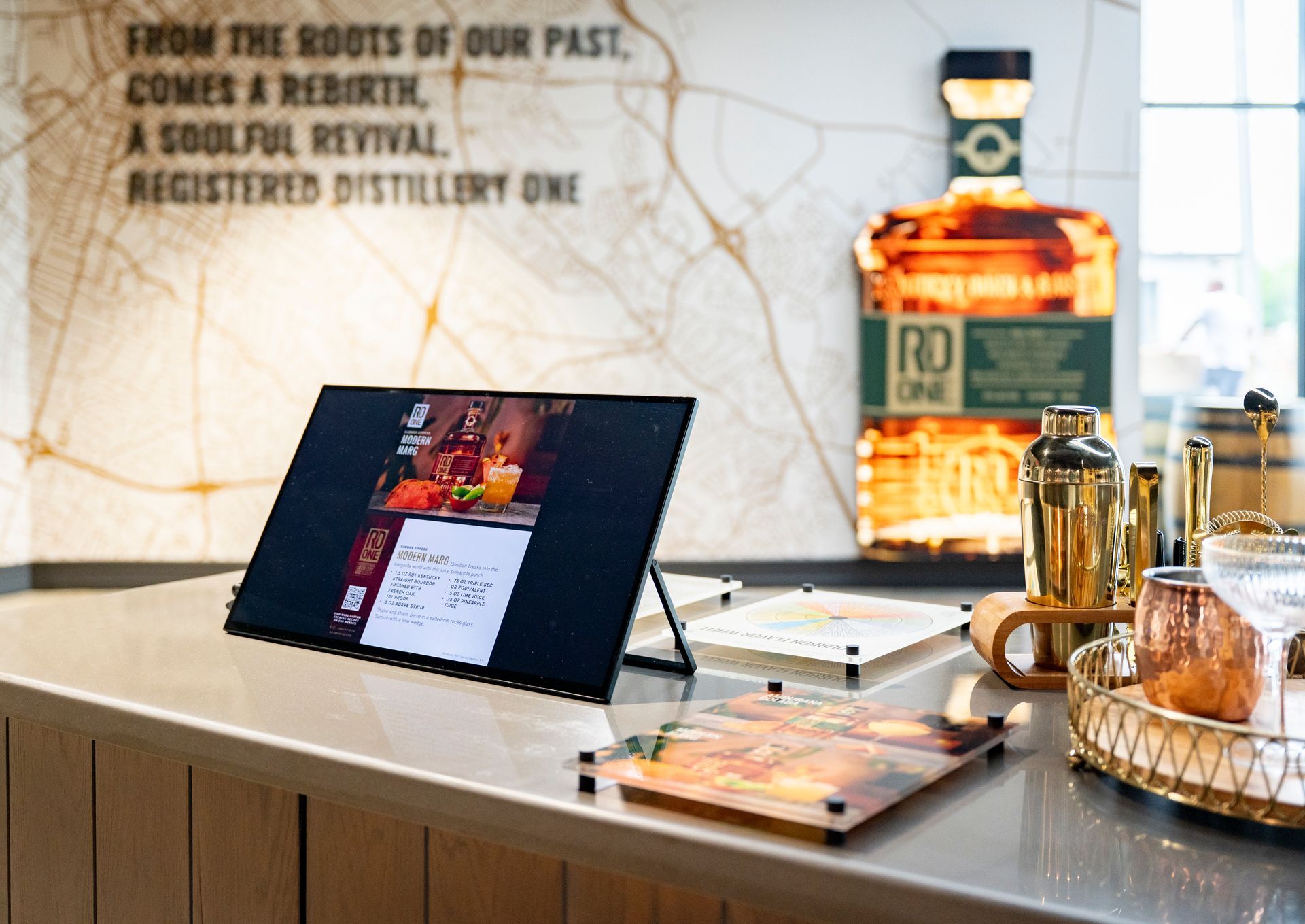 Bar with display screen, cocktail supplies, and a large illuminated bottle outline, text overlay.