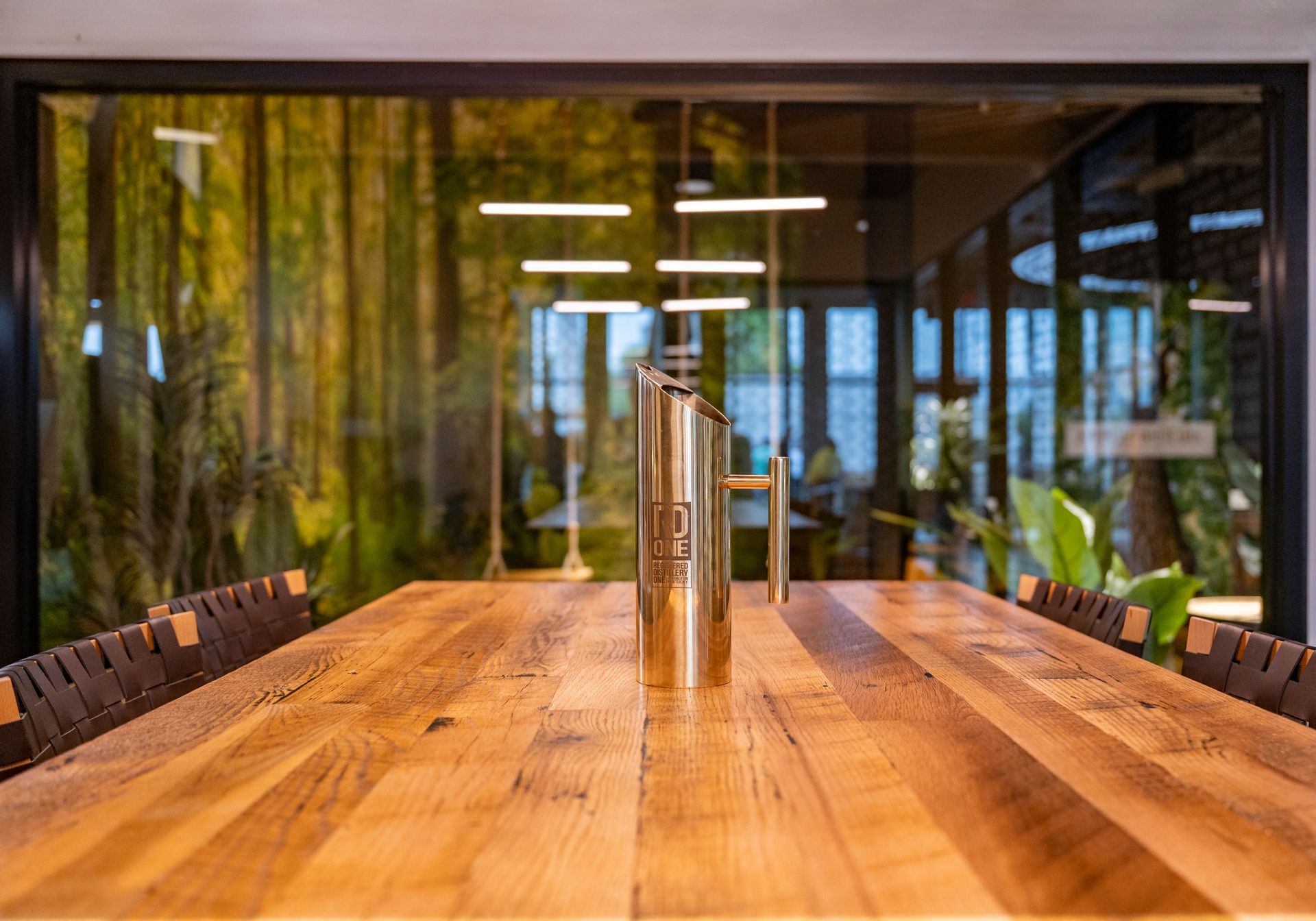 Wooden table with a metallic pitcher, set against a glass wall with a forest mural and overhead lights.