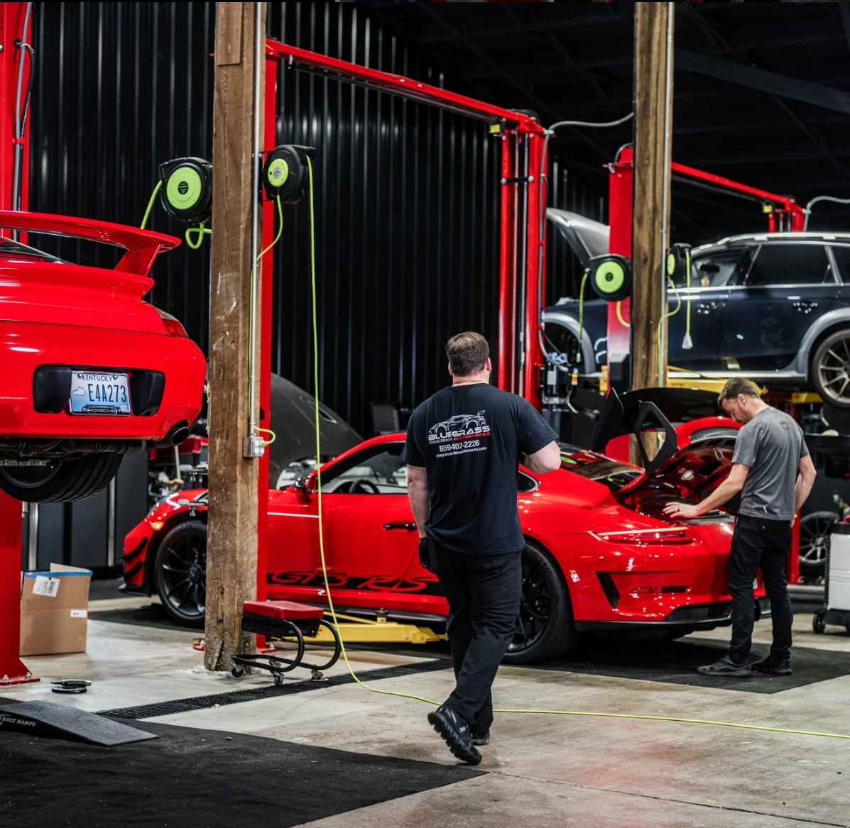 Two mechanics working on red sports cars in a garage with lifts; one walks, one looks at engine.