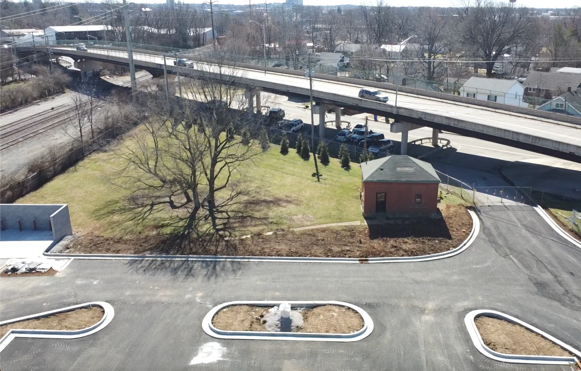 Overhead view of a highway overpass, parking lot, small brick building, and bare trees under a bright sky.