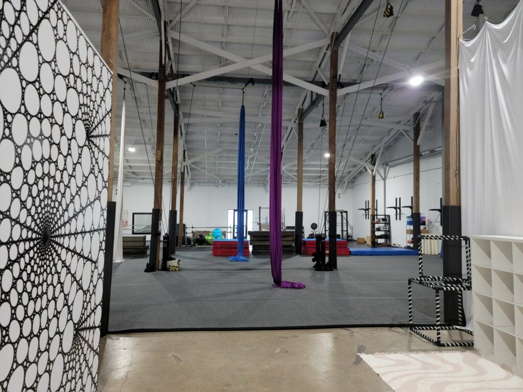 An open warehouse with aerial silks hanging; a black and white patterned art piece is on the left.