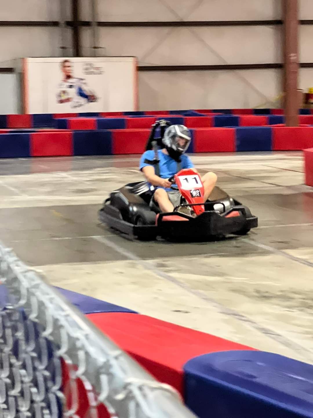 Person driving a black go-kart on an indoor track, wearing a helmet. Red and blue track barriers.