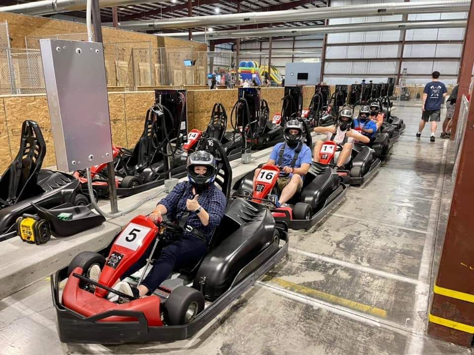 People in go-karts lined up indoors, wearing helmets, ready to race.