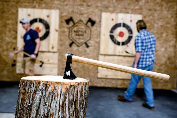 Axe on a wood stump, two men throwing axes at targets in an axe-throwing range.