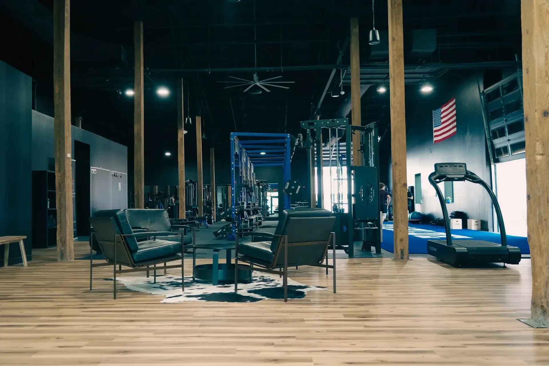 Interior of a gym with exercise equipment, seating area, American flag, and wooden columns.