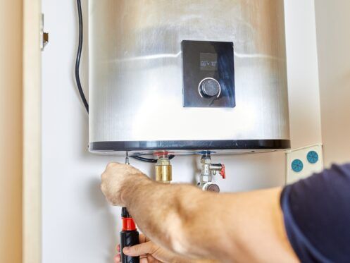 Person installing a water heater, using a screwdriver on a fitting. Silver tank.