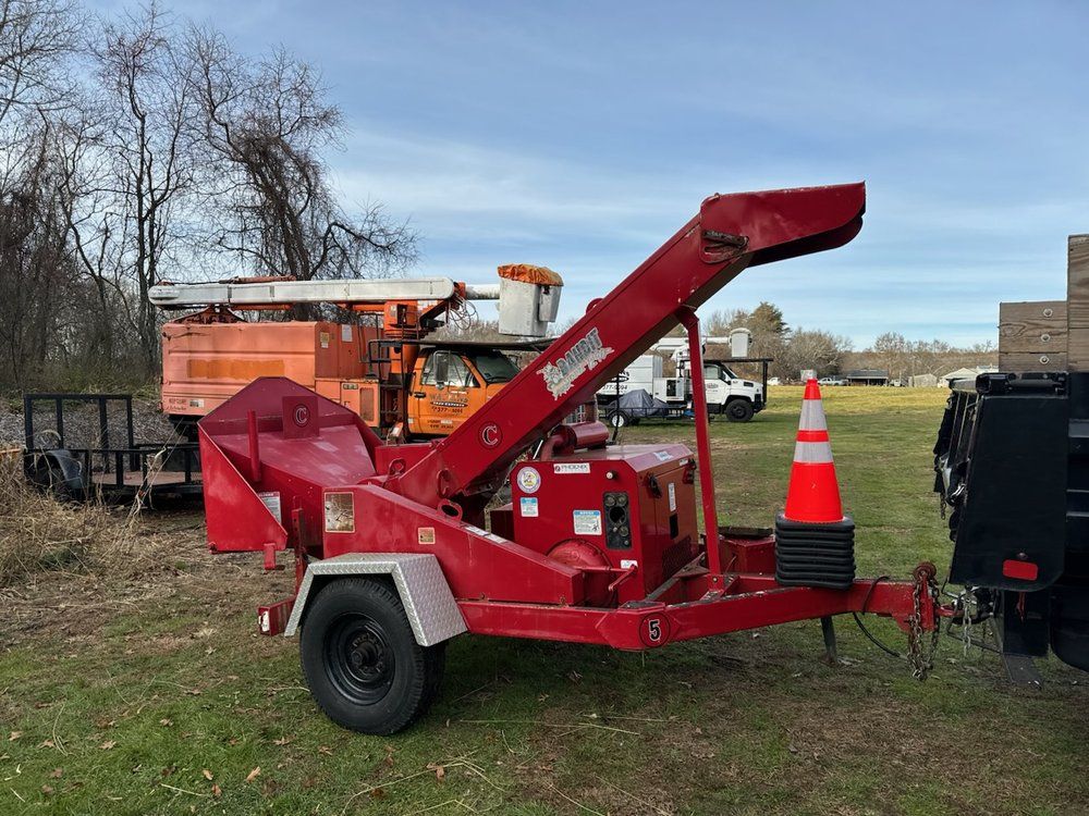 Red wood chipper on a trailer, parked on grass. Orange cone in front, with utility trucks in the background.