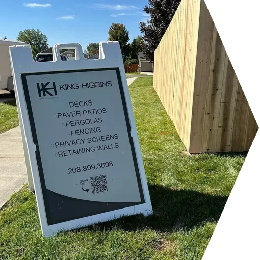 A sidewalk sign for King Higgins landscaping stands on grass beside a wooden structure.