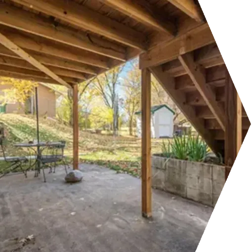 Covered patio beneath wooden deck with stairs, concrete floor, table, and small yard in the background