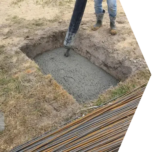 Worker pouring concrete into a dug-out ground hole beside stacked rebar.
