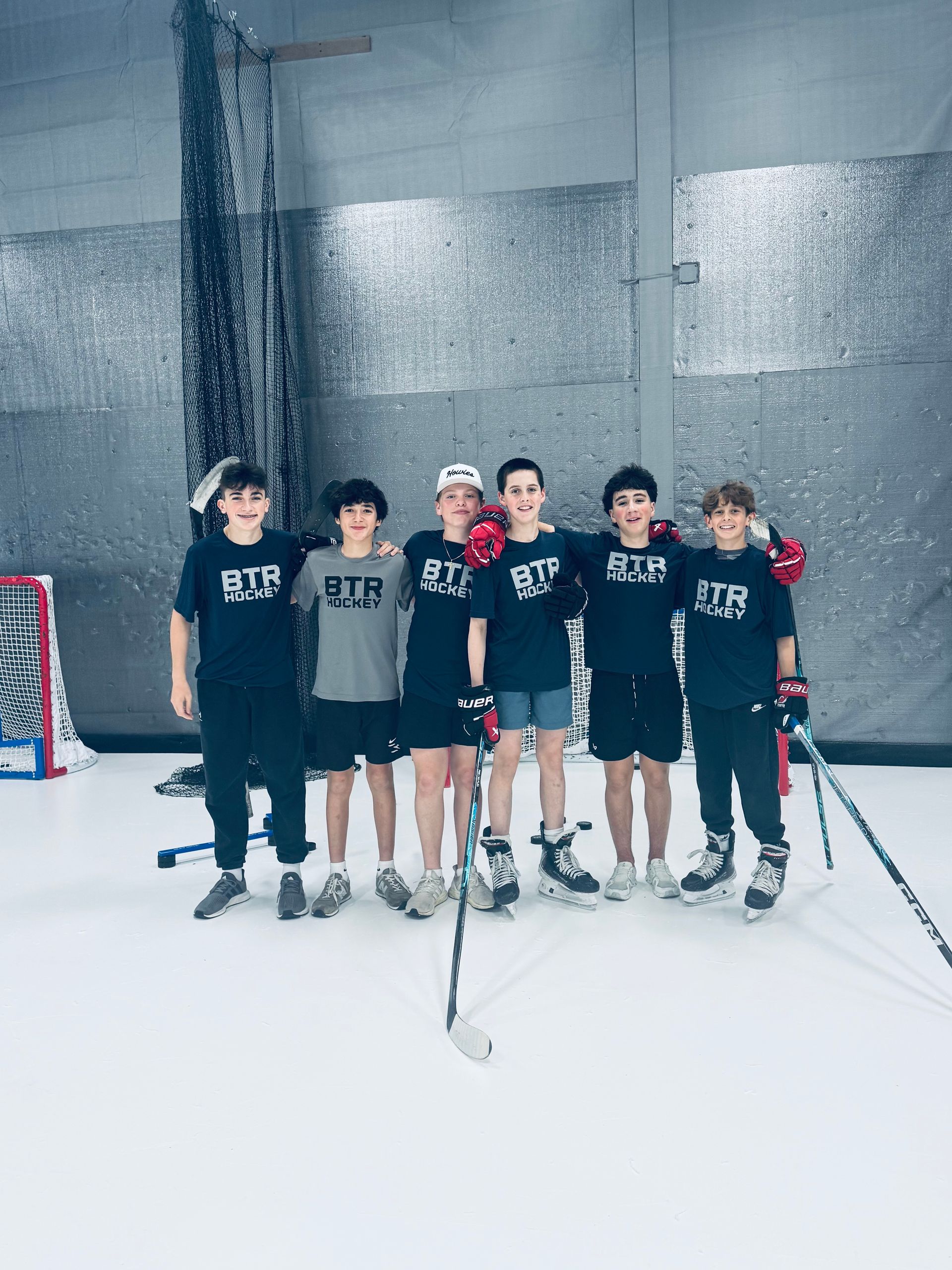 Six youths posing on an indoor hockey rink in dark jerseys, holding sticks, with a goal nearby.