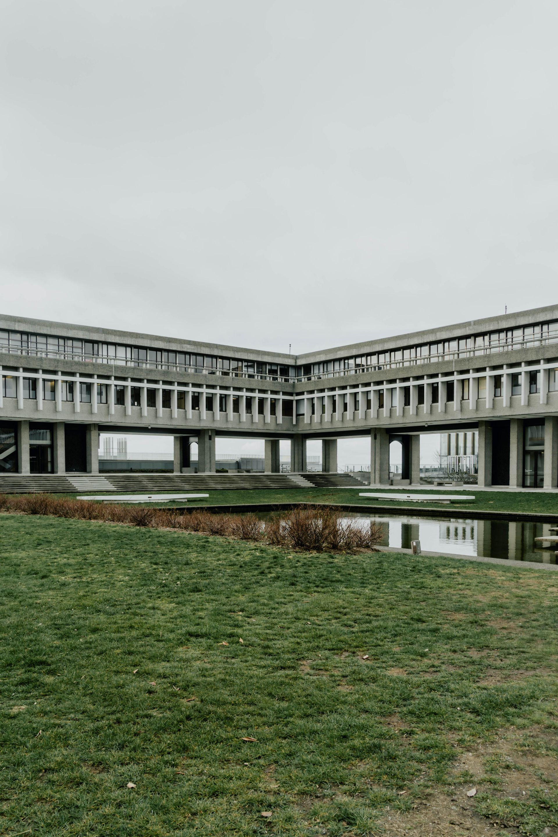 Modern concrete building with a long covered walkway reflected in a shallow pool, under cloudy skies