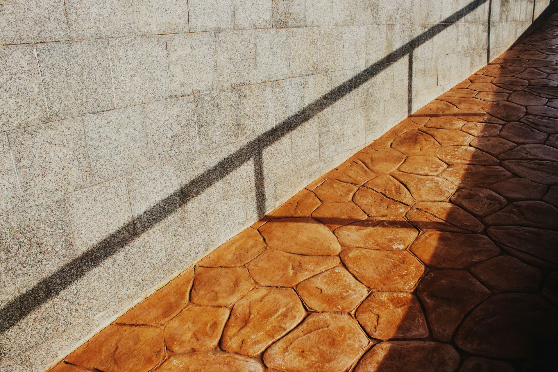 Sunlit stone walkway beside a textured wall, with a sharp diagonal shadow across orange paving stones