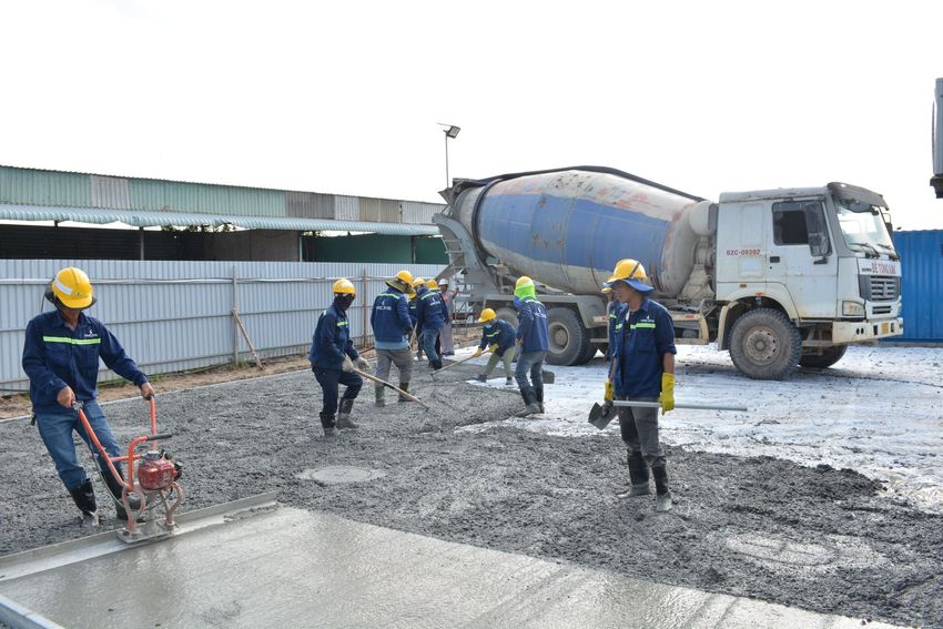 Construction workers in yellow hard hats paving wet concrete beside a concrete mixer truck