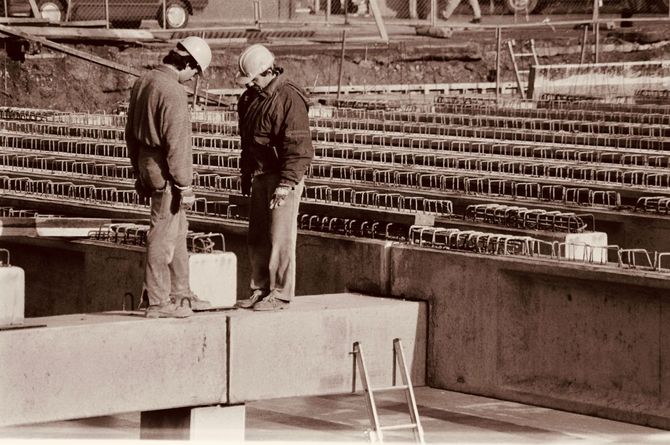 Two workers in hard hats standing in a stadium under construction, with rows of seats behind them.
