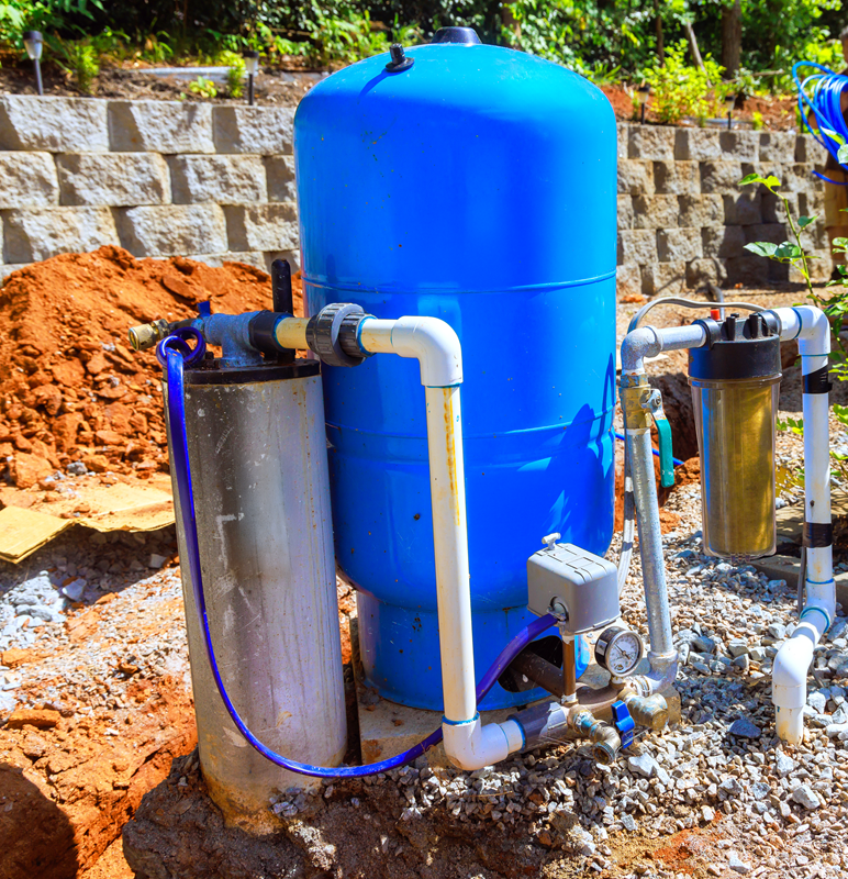 Blue water filtration tank and pumps installed outdoors on gravel near a stone retaining wall.