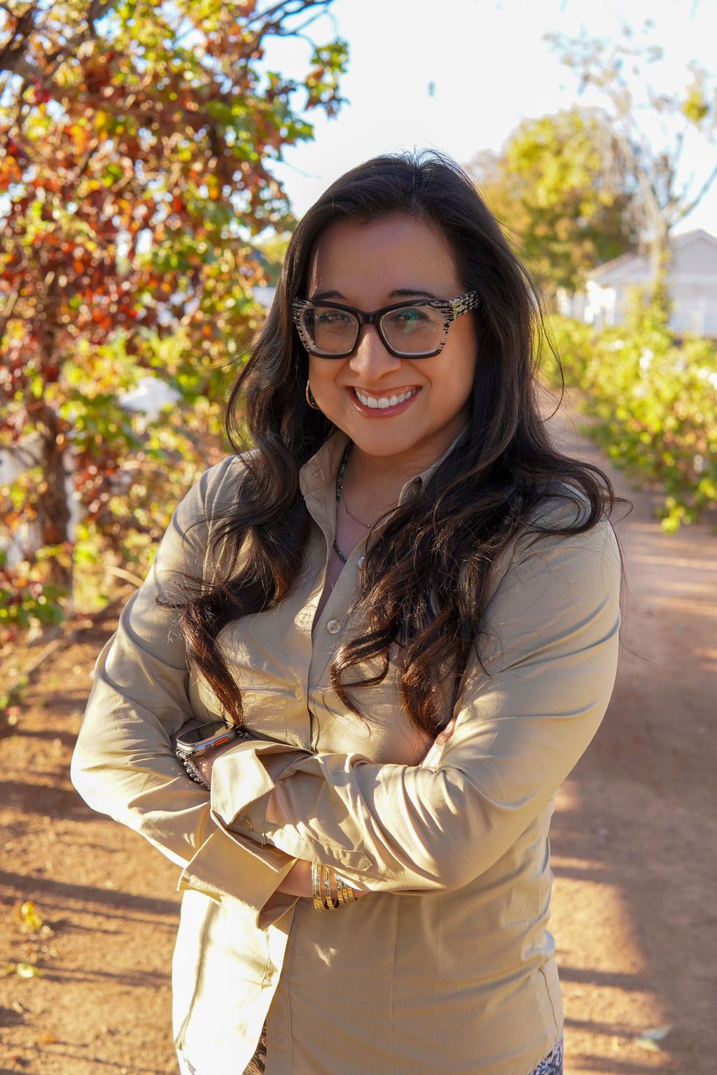 A woman wearing glasses and a tan jacket is standing with her arms crossed and smiling.