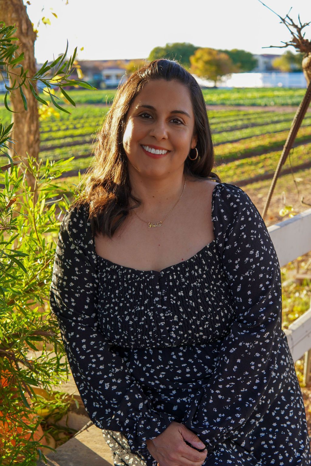 A woman in a black dress is standing next to a tree and smiling.