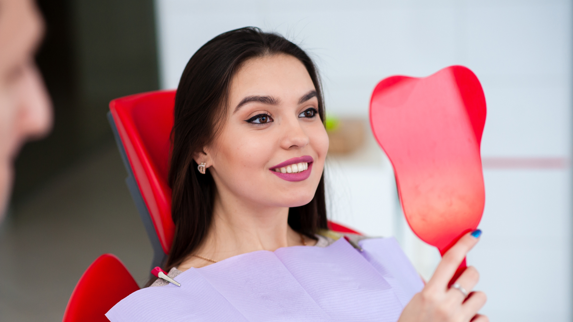 A woman is sitting in a dental chair looking at her teeth in a mirror.