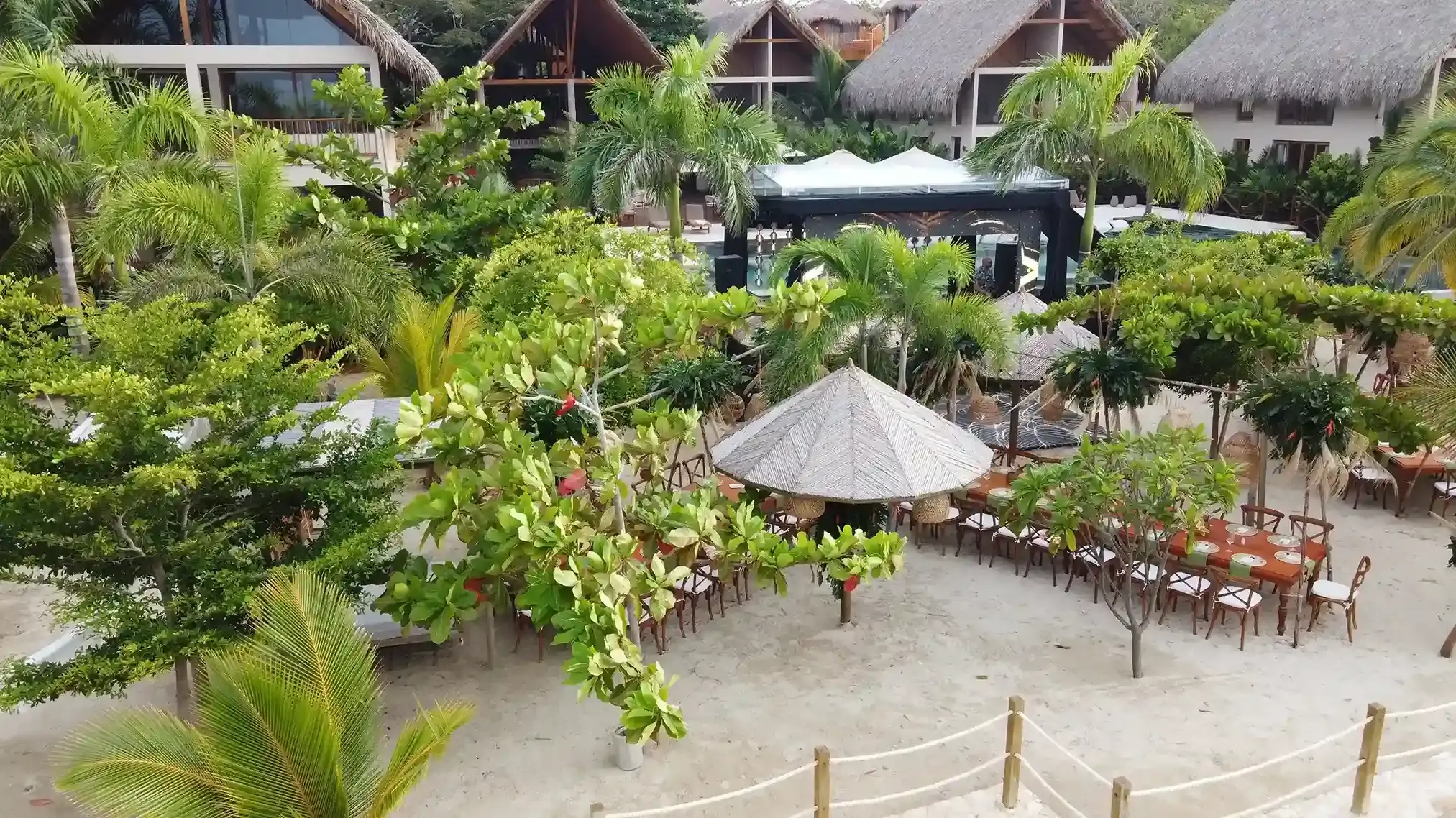 Montagem de mesas para casamento na praia sob quiosques de palha e palmeiras no Hotel Aimarawa