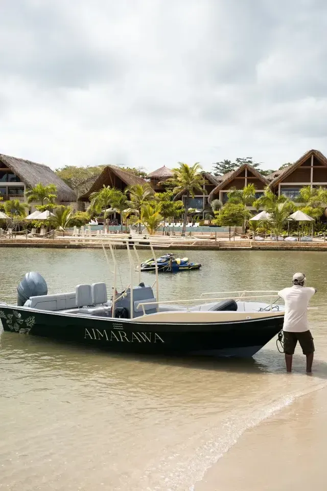 Lancha de Aimarawa en la orilla, con cabañas, palmeras y piscina de hotel frente a la playa