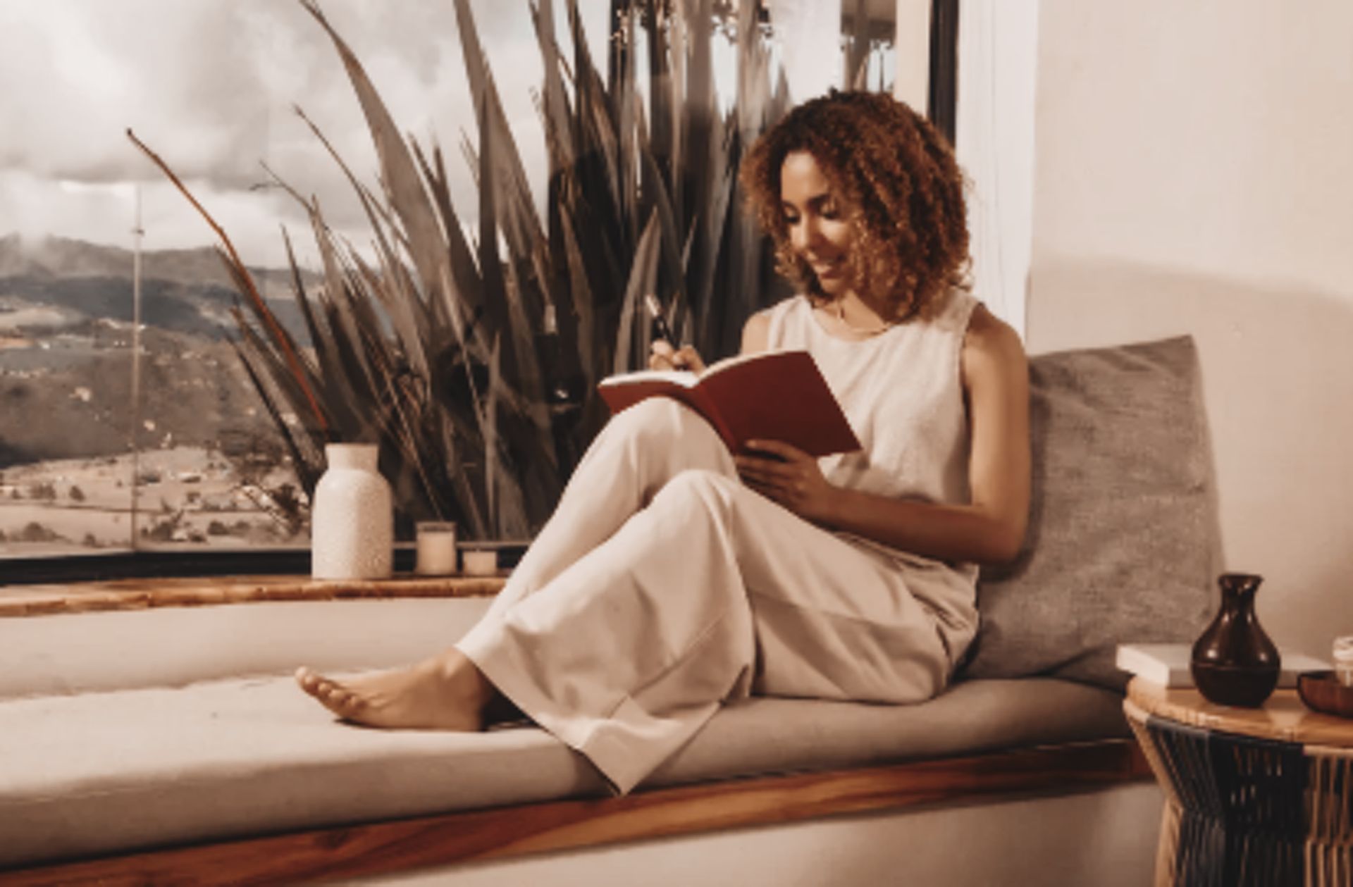 A woman writing thoughtfully in a journal near a wide window