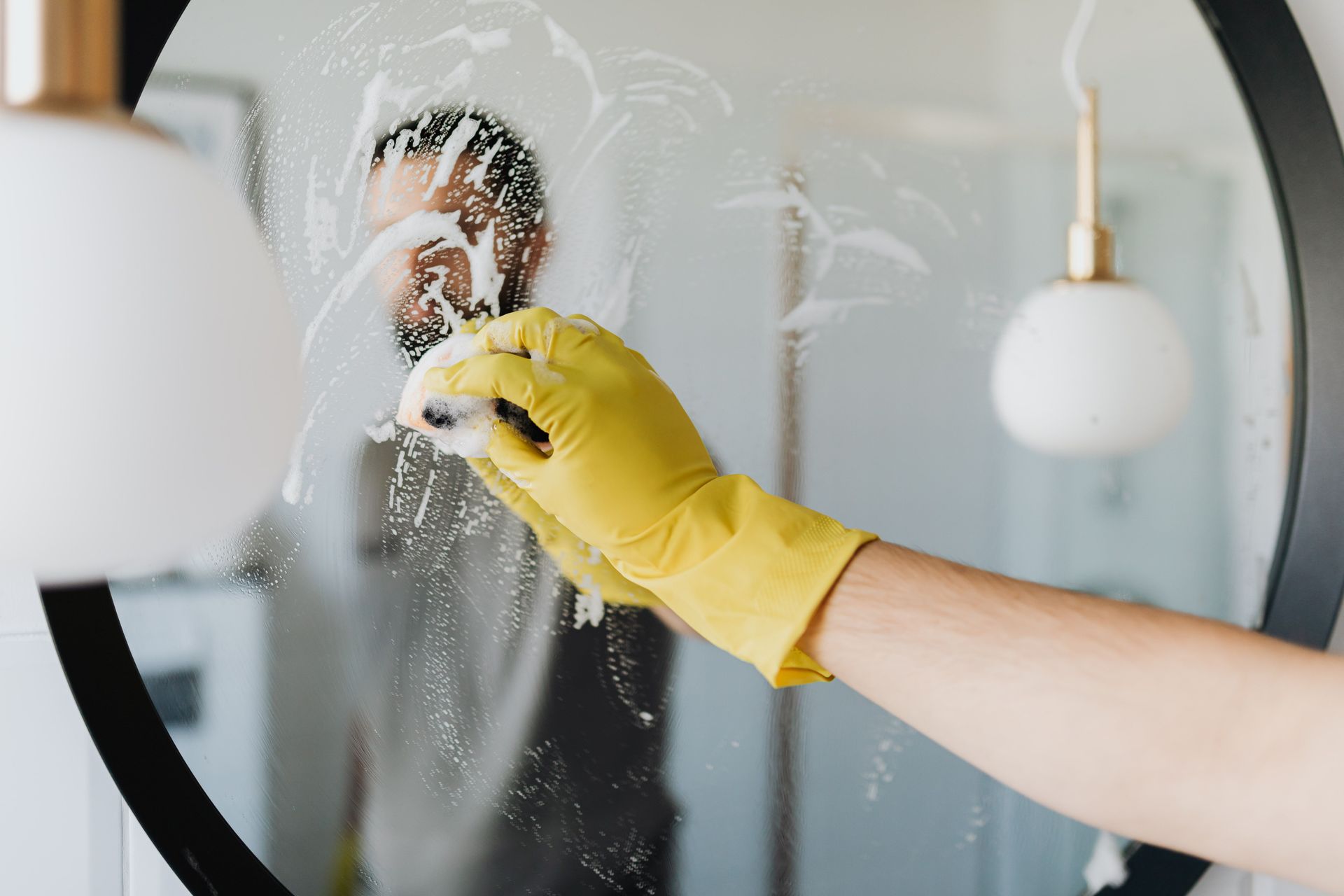 A hand in a yellow glove wiping a soapy bathroom mirror.