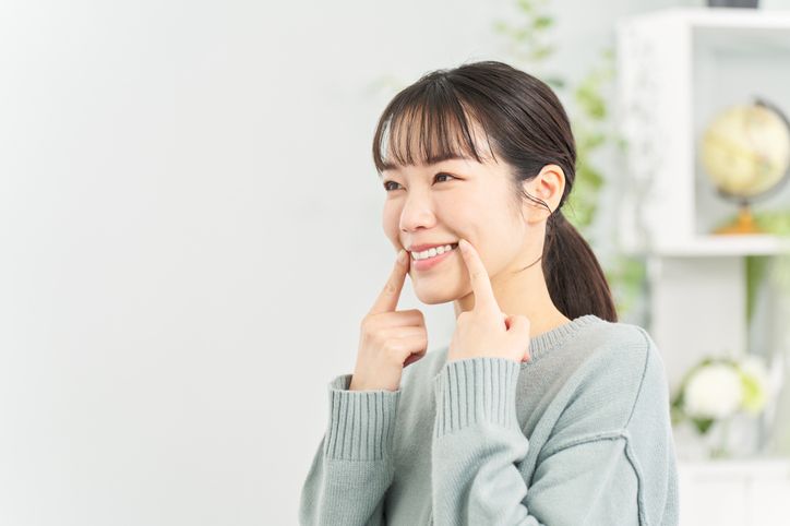 Woman smiling, pointing to her cheeks, indoors.
