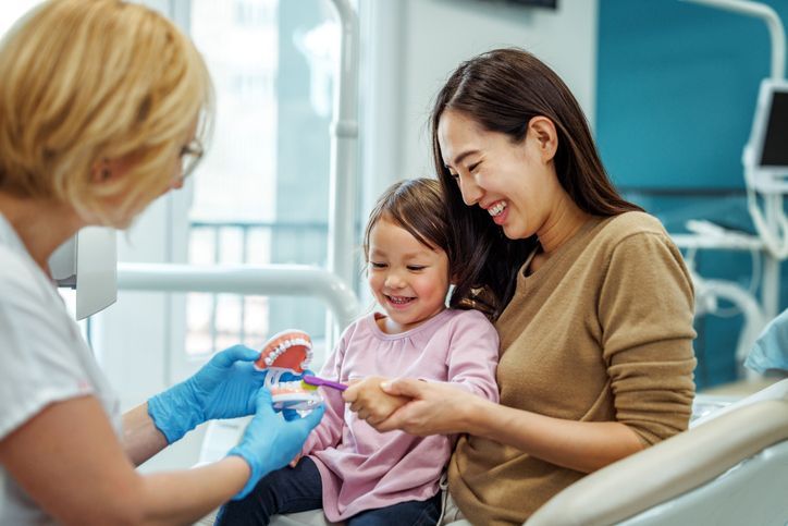 Dentist showing a young child how to brush teeth on a model, with the child's mother present.