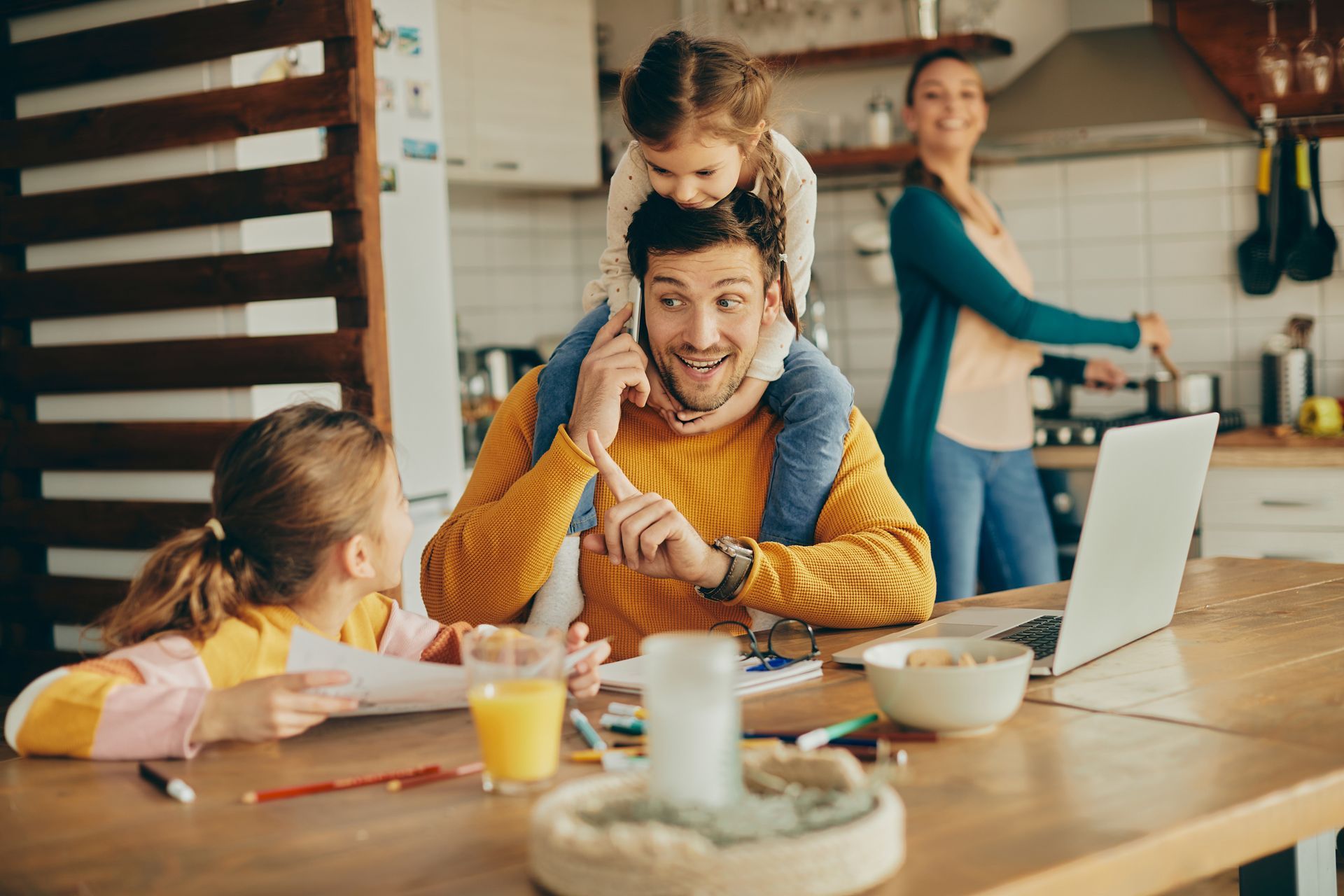 Man with child on shoulders, working from home at kitchen table with two other children and woman cooking.