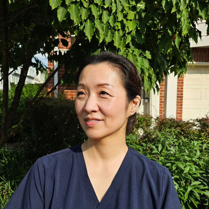 Ellie in a navy top standing outdoors under leafy trees, looking slightly to the side and smiling.
