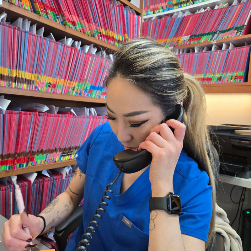 Esther in blue scrubs writing while speaking on a desk phone in a file-filled office