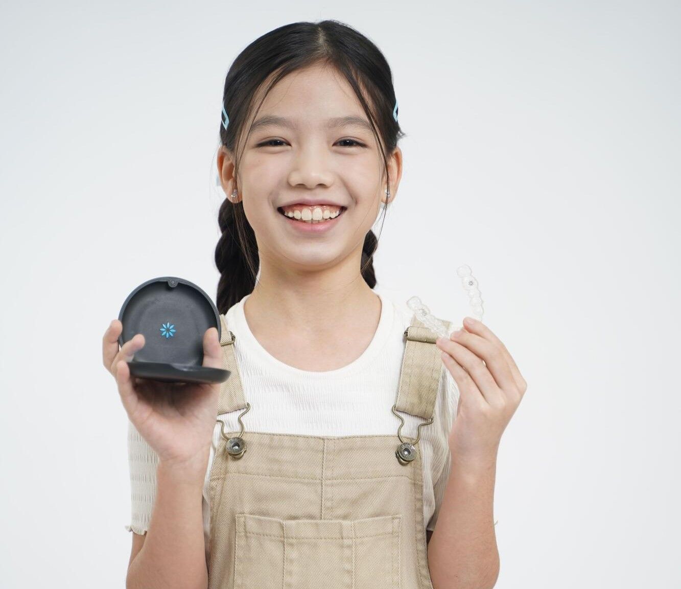 Smiling person in beige overalls holding a black device and a small white item against a light background