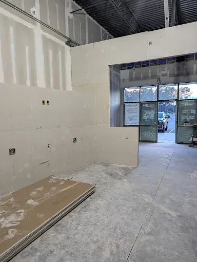 An unfinished interior space under construction with drywall panels on walls and a view of an outdoor parking lot.