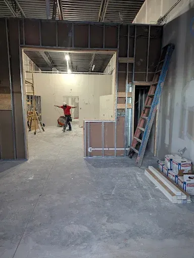 A construction site with a worker in a red shirt moving a bucket through a doorway, with a tall ladder leaning nearby.