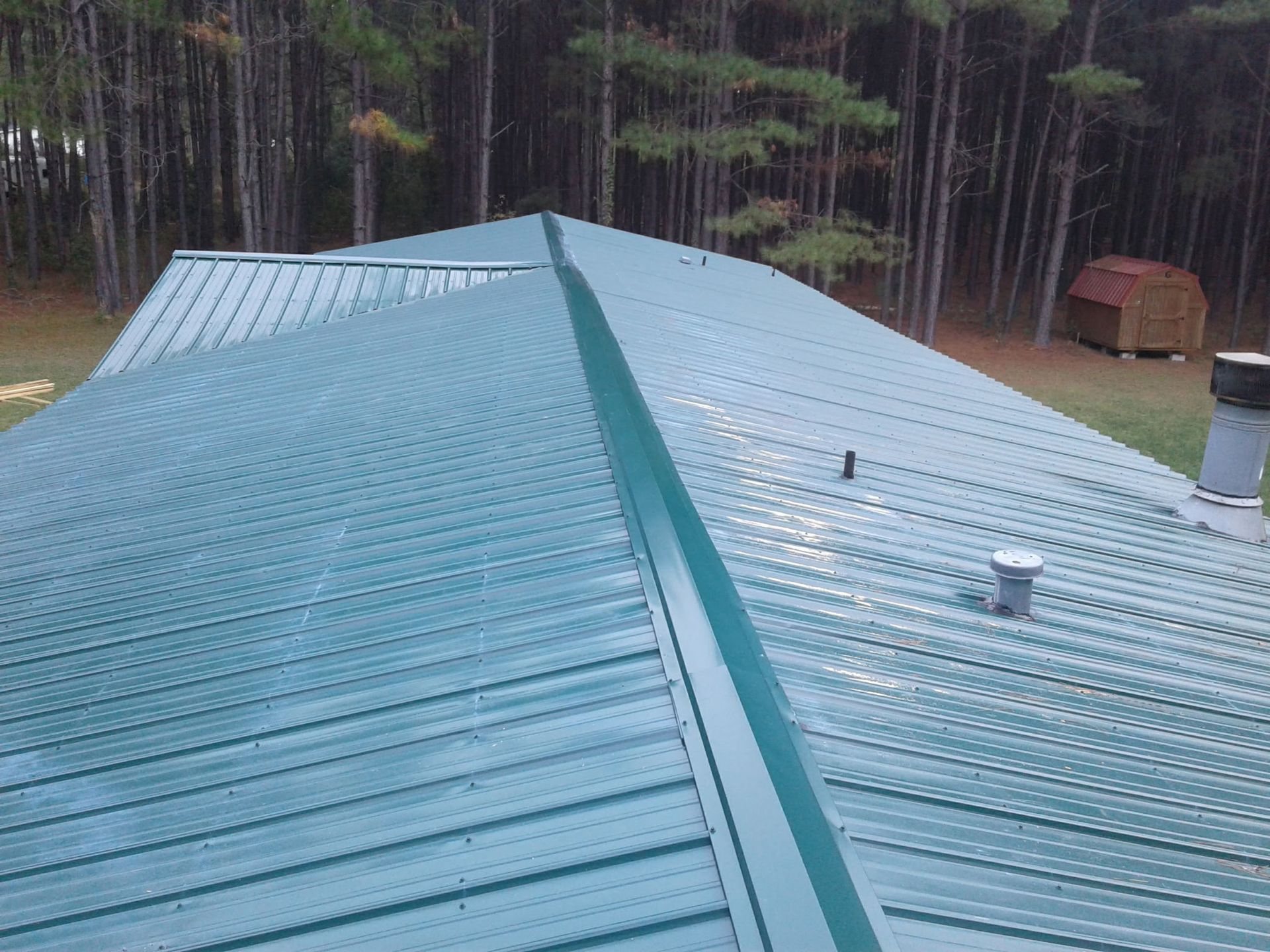 A green metal roof featuring a ridge cap and damaged flashing, set against a backdrop of pine trees and a small shed.