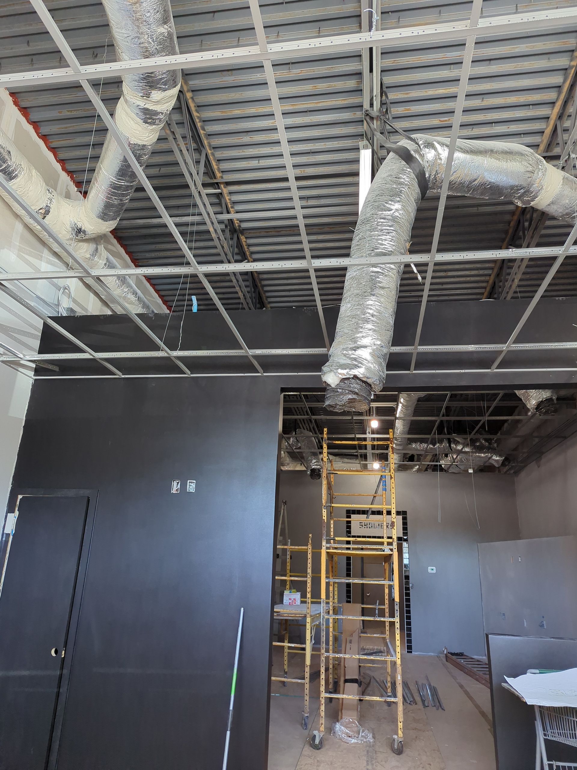 Interior construction site with exposed metal ceiling grid, silver ductwork, black wall, and yellow scaffolding.