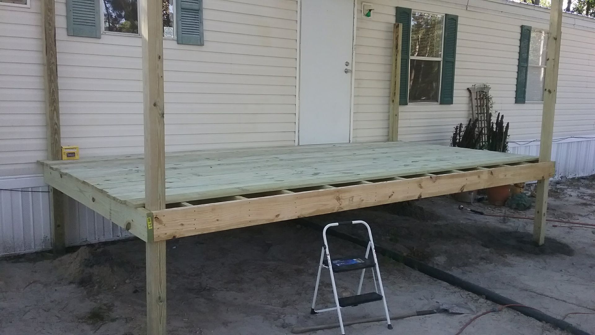 A newly constructed wooden deck attached to a light-colored mobile home, with a small stepladder standing on the dirt below.