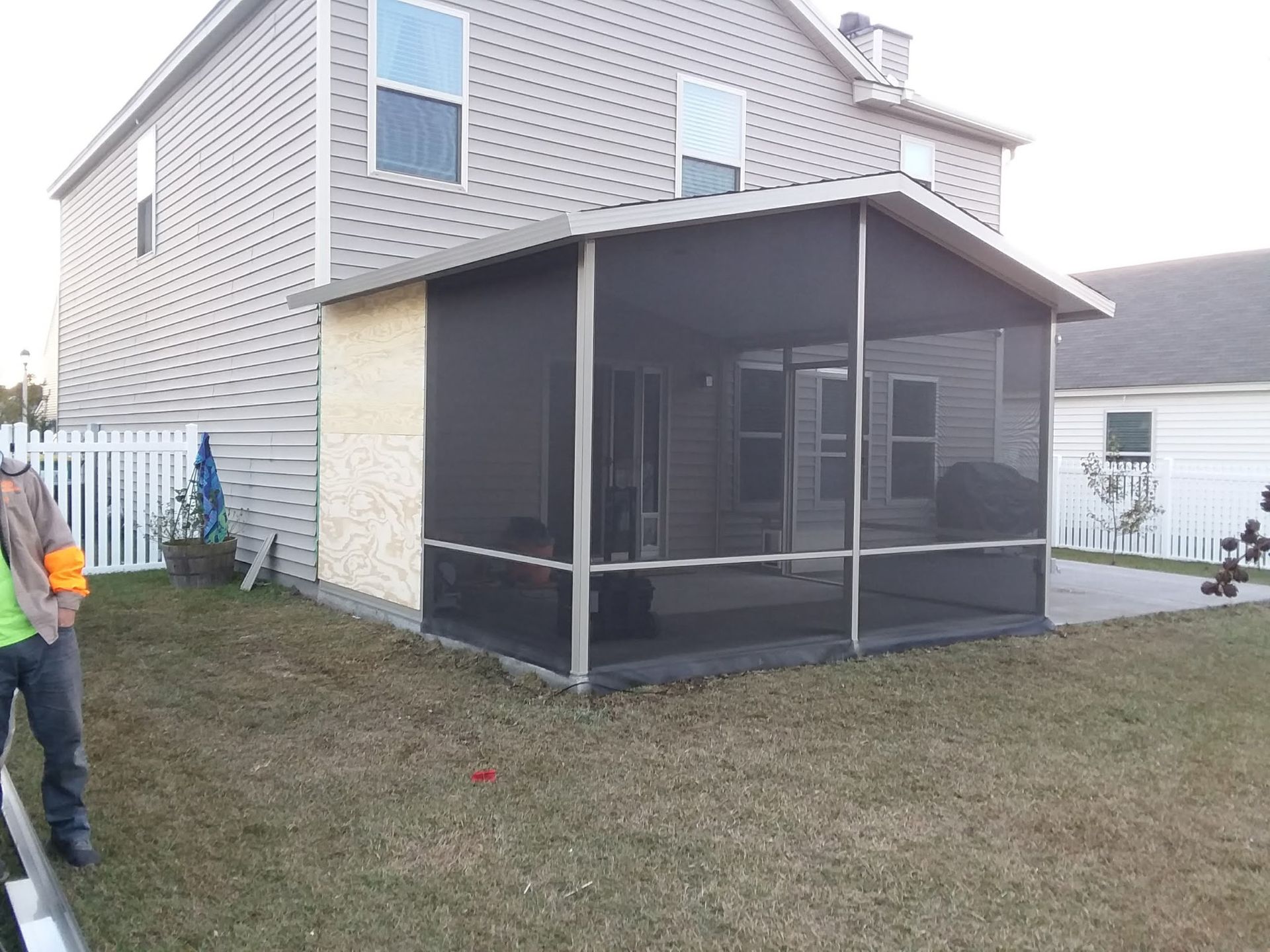 A tan two-story house with a screened-in porch and a partial plywood exterior wall in a residential backyard.