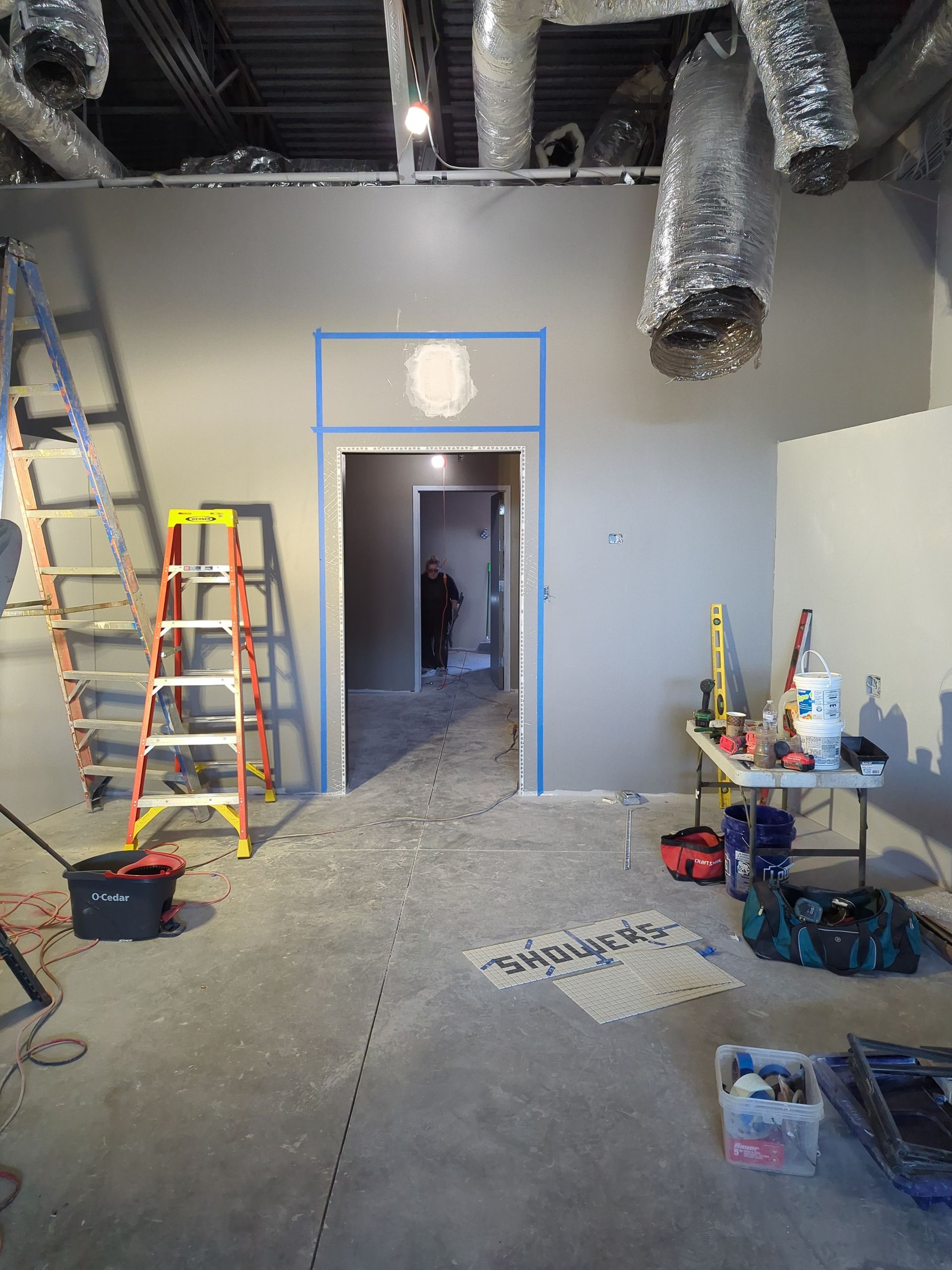 A construction site showing a doorway with blue painter's tape, two step ladders, tools, and exposed ceiling ductwork.