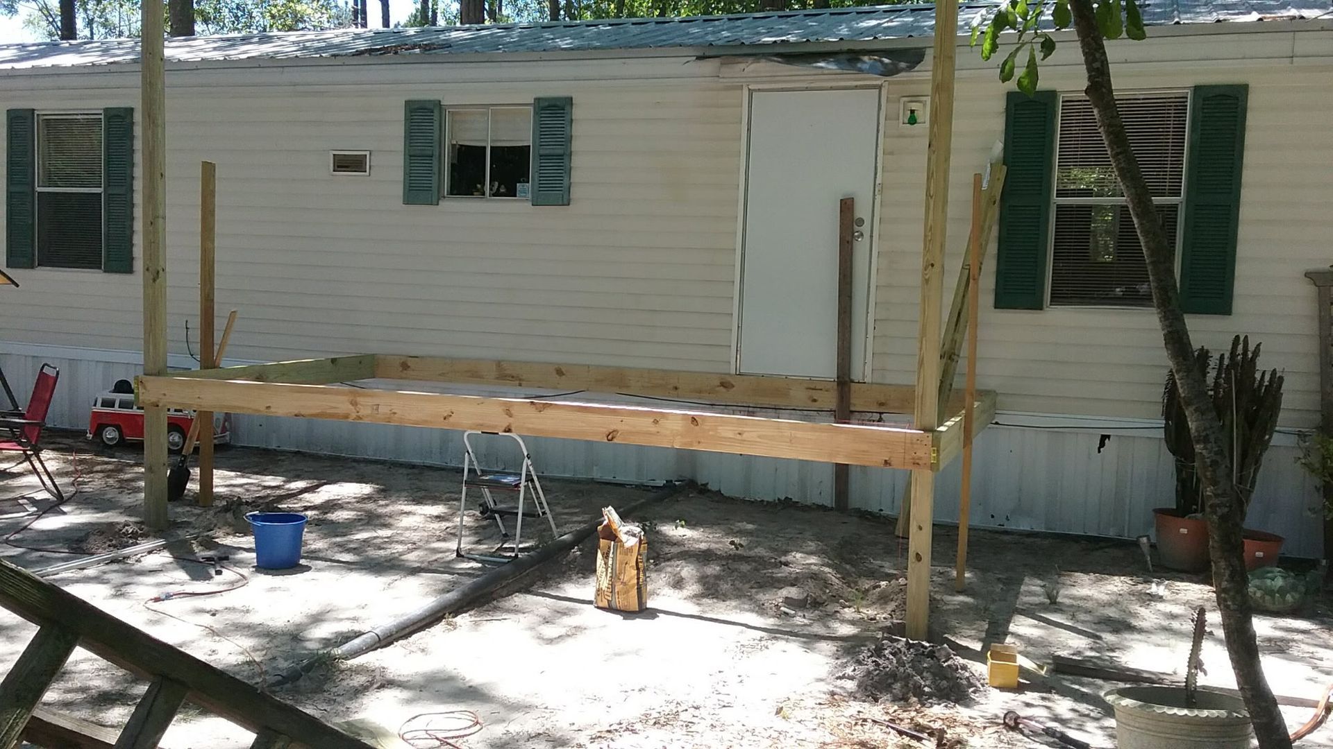 A wooden deck frame under construction in front of a light-colored mobile home, supported by vertical posts.